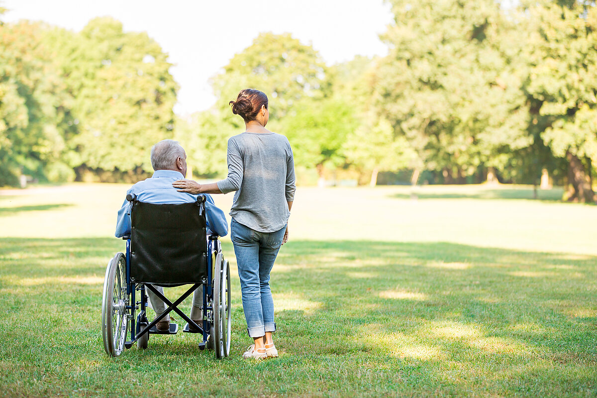 Senior im Rollstuhl und neben ihm steht eine Pflegerin.