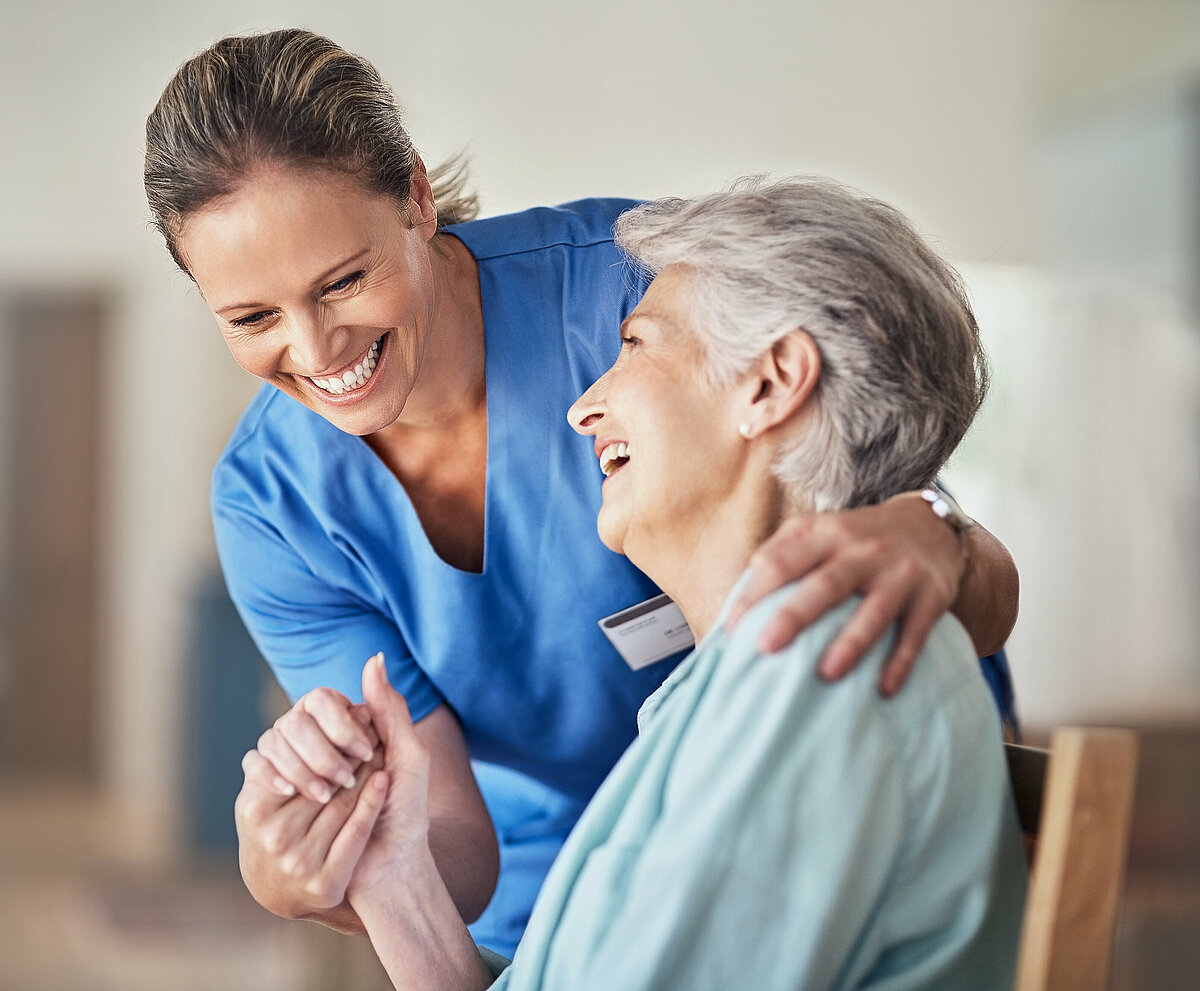 Pflegerin und Seniorin halten sich an der Hand während sie gemeinsam lachen.