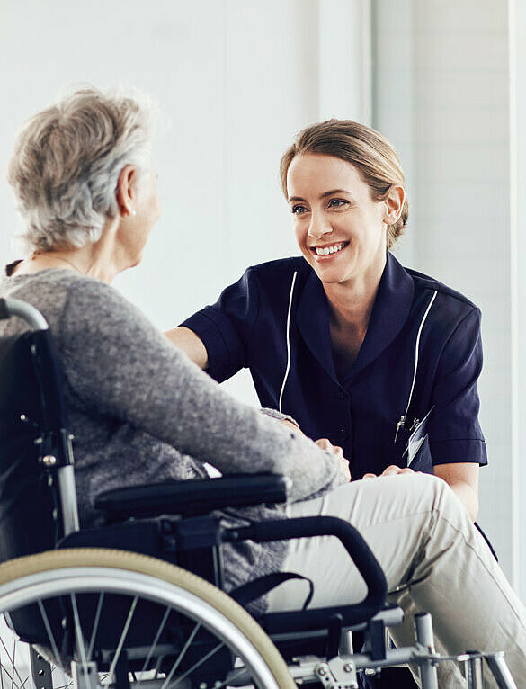 Pflegerin mit Weiterqualifizierung bei Alloheim beugt sich runter zu Seniorin im Rollstuhl